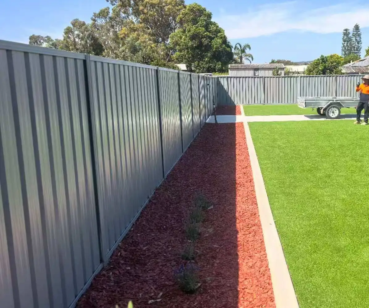Grey Colorbond boundary fence running along a backyard, with a red-mulch garden bed at its base