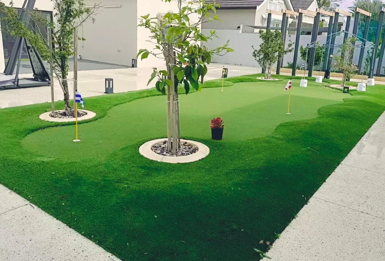 Artificial grass putting green with holes and flags, and feature trees set into circular stone-ringed planters